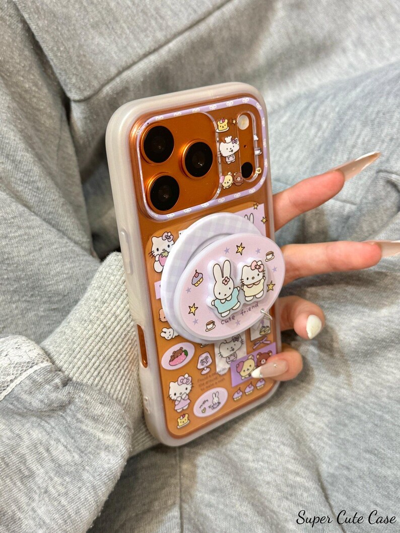 May include: A clear phone case with a gold-colored interior and a pop-up grip featuring cartoon characters. The case has a white border and is decorated with various cartoon images, including Hello Kitty and a bunny. The text "Super Cute Case" is visible.