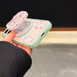 May include: A clear phone case with a pink and mint green design featuring cartoon bunnies and strawberries. The case has a pop-up grip with a pink bow and a cartoon cat face. The words "Super Cute Case" are visible in the bottom right corner.
