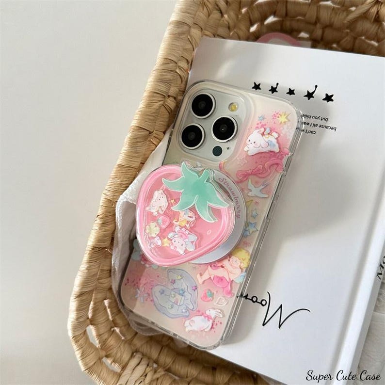 May include: Clear phone case with a pink strawberry-shaped phone grip. The case features a whimsical design with pink, white, and blue illustrations of angels, bunnies, and stars. The phone case is inside a woven basket.