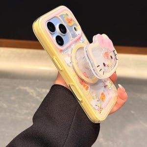 Peut inclure: Coque de t&eacute;l&eacute;phone jaune avec un dos transparent orn&eacute; d'un motif de dessin anim&eacute; et d'une poign&eacute;e pop-up. La poign&eacute;e pop-up a la forme d'un personnage avec un n&oelig;ud rose. La coque a une bordure jaune et un dos transparent avec des graphiques de dessins anim&eacute;s.