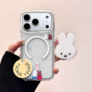 May include: Clear phone case with a white magnetic ring and a white bunny-shaped phone grip. The case has a tag that says "Y2CASE". The case has a small white, red, and blue bunny design. The phone grip has a black cross for a mouth.