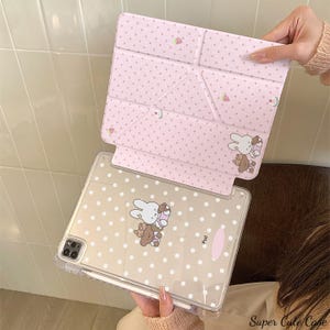 Könnte beinhalten: Eine rosa iPad-Hülle mit passendem Cover, beide mit weißen Punkten und Cartoon-Hasen- und Bären-Illustrationen verziert. Das Cover lässt sich zu einem Ständer zusammenfalten. Die Hülle ist transparent mit einem Stifthalter.