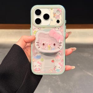 May include: A clear phone case with a mint green border featuring a Hello Kitty design. The case has a 3D Hello Kitty face with a pink bow and a pop-up feature. The background is filled with Hello Kitty characters and other cute graphics.