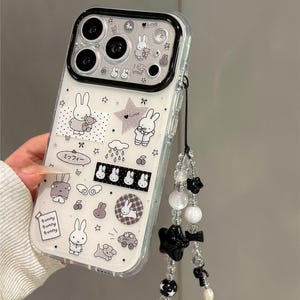 Pode incluir: Capa de telem&oacute;vel transparente com borda preta, com desenhos de coelhos brancos e estrelas. Inclui uma al&ccedil;a de pulso com contas e detalhes de estrelas e la&ccedil;os pretos. O texto "Bunny Bunny Bunny" &eacute; vis&iacute;vel.