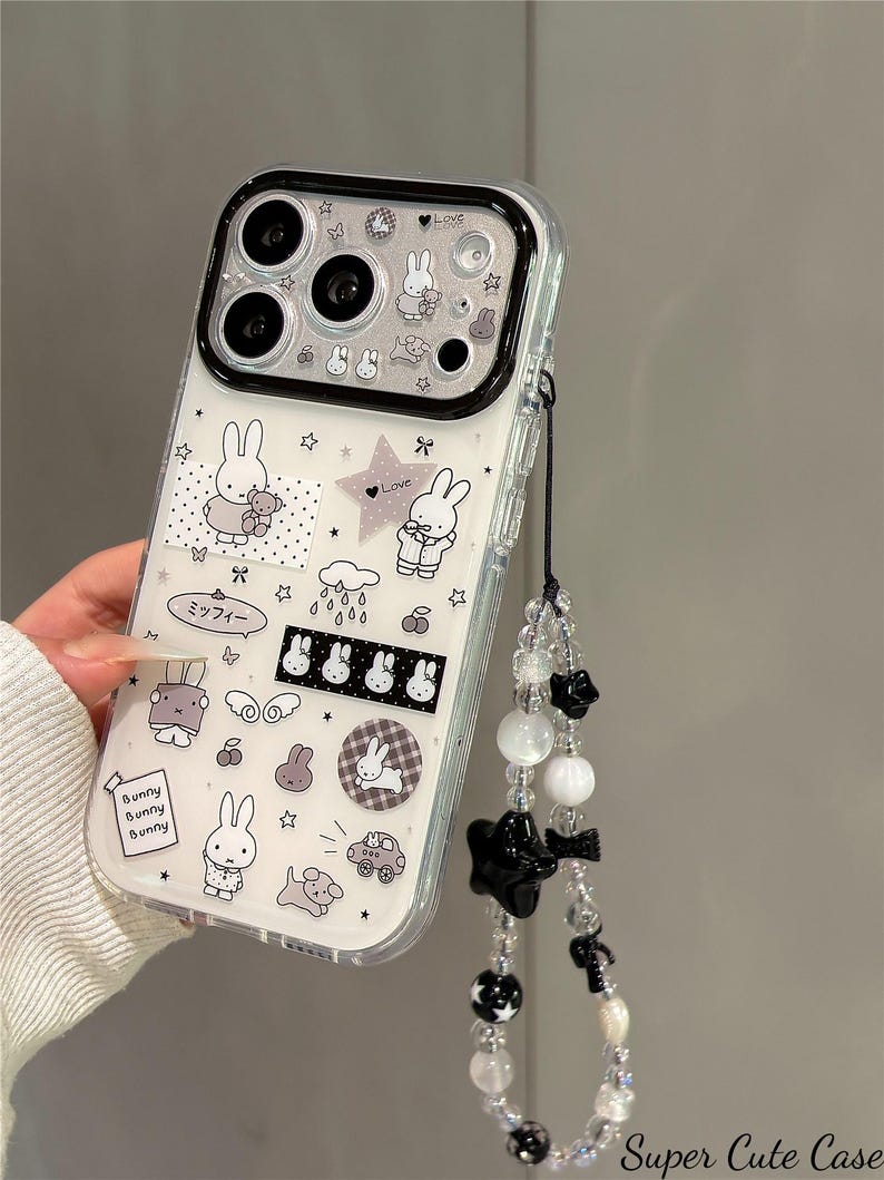 Pode incluir: Capa de telem&oacute;vel transparente com borda preta, com desenhos de coelhos brancos e estrelas. Inclui uma al&ccedil;a de pulso com contas e detalhes de estrelas e la&ccedil;os pretos. O texto "Bunny Bunny Bunny" &eacute; vis&iacute;vel.