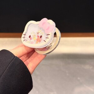 Peut inclure: Un &eacute;tui compact et transparent avec un design Hello Kitty, un n&oelig;ud rose et une surface miroir. L'&eacute;tui a une bordure blanche et contient de petites figurines color&eacute;es. Le texte "Super Cute Case" est visible.