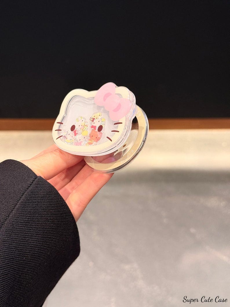 Peut inclure: Un &eacute;tui compact et transparent avec un design Hello Kitty, un n&oelig;ud rose et une surface miroir. L'&eacute;tui a une bordure blanche et contient de petites figurines color&eacute;es. Le texte "Super Cute Case" est visible.