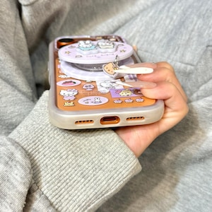 May include: A clear phone case with a cartoon design featuring a variety of characters and shapes in orange, pink, and white. The case has a pop-up feature and a charm. The text "Super Cute Case" is visible at the bottom.