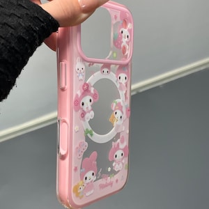 May include: Pink phone case with a clear center and a design featuring cartoon characters, strawberries, and stars. The case has a pink border and the words "Love Melody" are printed on the front. The case is held by a hand.