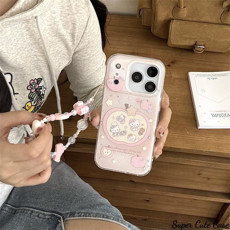 May include: A pink phone case with a Hello Kitty design, featuring apples and stars. The case has a clear strap with pink and white beads. The text "Super Cute Case" is visible at the bottom.