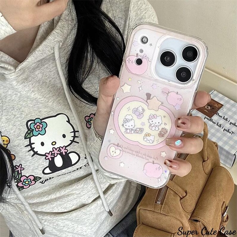 May include: Clear phone case with a pink apple design featuring Hello Kitty characters and stars. The case has a clear border and a pink background with a light pink apple shape. The phone case is held by a person.