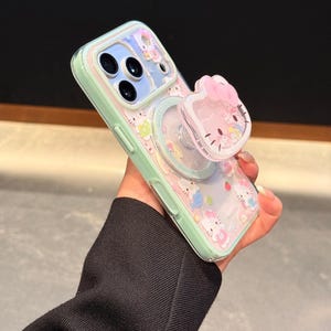 May include: A mint green phone case with a clear back featuring a repeating pattern of cartoon characters. The case has a matching pop-up grip with a pink bow and a cartoon face. The text "Super Cute Case" is visible.