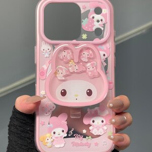 May include: Pink phone case featuring a My Melody design. The case has a clear back with pink accents and a raised My Melody character. Additional character graphics and the text "my Melody" are also visible.