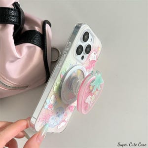 May include: Clear phone case with a colorful design featuring cartoon characters and a pink strawberry-shaped phone grip. The case has a clear back and a silver frame. The text "Super Cute Case" is visible in the bottom right corner.