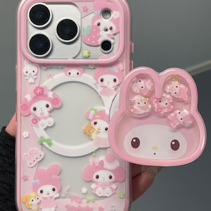 Puede incluir: Funda de teléfono rosa con parte trasera transparente y pop-socket a juego. La funda presenta personajes de My Melody y otros diseños bonitos. El pop-socket tiene un diseño de cara de My Melody y varios personajes de My Melody.