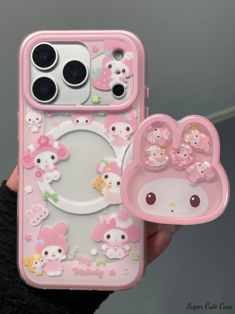 May include: Pink phone case with a clear back and a matching pop-socket. The case features My Melody characters and other cute designs. The pop-socket has a My Melody face design and several My Melody characters. The case is designed to fit a phone.
