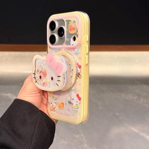 Peut inclure: Coque de t&eacute;l&eacute;phone jaune avec des personnages Hello Kitty et une poign&eacute;e pop-up. L'&eacute;tui a un dos transparent avec un bord jaune et un n&oelig;ud rose sur la poign&eacute;e pop-up. L'&eacute;tui est d&eacute;cor&eacute; de diverses illustrations Hello Kitty.
