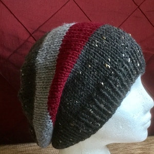 May include: A grey, red and black striped knitted beanie hat with a textured pattern. The hat is on a white mannequin head.