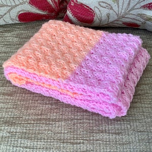 May include: A pink and peach crocheted baby blanket with a textured pattern.