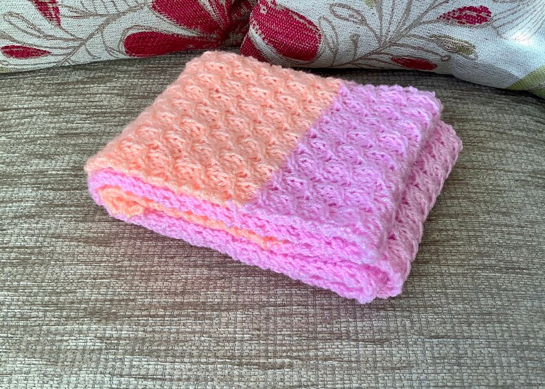 May include: A pink and peach crocheted baby blanket with a textured pattern.