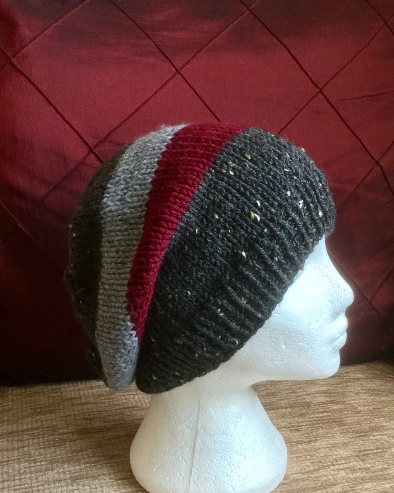 May include: A grey, red and black striped knitted beanie hat with a textured pattern. The hat is on a white mannequin head.