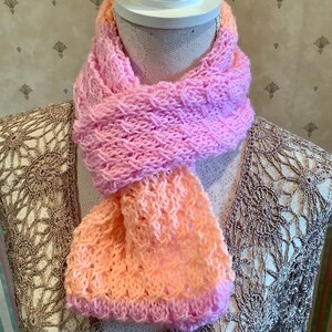May include: A pink and peach knitted scarf with a textured pattern. The scarf is displayed on a white mannequin.