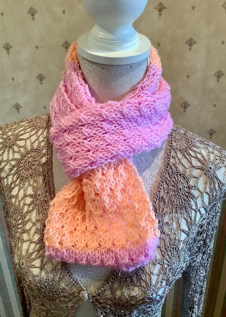 May include: A pink and peach knitted scarf with a textured pattern. The scarf is displayed on a white mannequin.