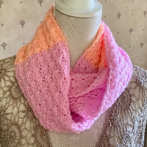 May include: A pink and peach knitted infinity scarf with a delicate, openwork pattern. The scarf is soft and lightweight, perfect for layering in the cooler months.