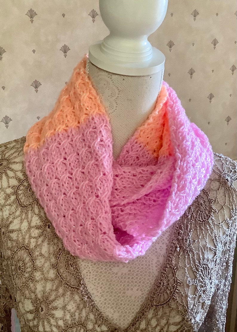 May include: A pink and peach knitted infinity scarf with a delicate, openwork pattern. The scarf is soft and lightweight, perfect for layering in the cooler months.