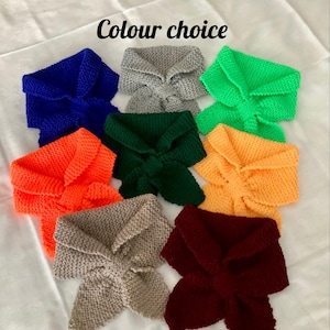 May include: A selection of eight knitted neck warmers in various colours, including blue, grey, green, orange, beige, and burgundy. The neck warmers are arranged in a semi-circle on a white surface. The text "Colour choice" is written in black above the neck warmers.