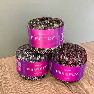 May include: Three spools of Sirdar Firefly yarn in various colors, with metallic threads. Each spool has a purple label with the brand name and product name. The yarn is on a wooden surface.