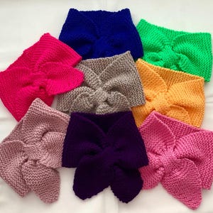 May include: A collection of eight knitted headbands featuring a central twist design. The headbands are available in a variety of solid colors including bright pink, royal blue, neon green, beige, orange, dusty rose, deep purple, and light pink.