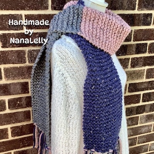 May include: A chunky knit scarf featuring color-blocked sections in dusty pink, dark gray, and deep navy blue with a speckled texture. The scarf includes fringe detailing at the ends and displays the text Handmade by NanaLelly on the left side.