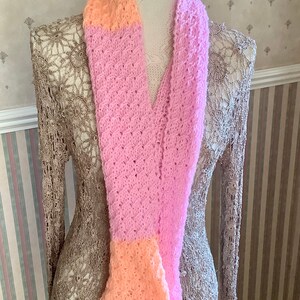 May include: A pink and peach knitted scarf with a lacy pattern. The scarf is displayed on a mannequin.