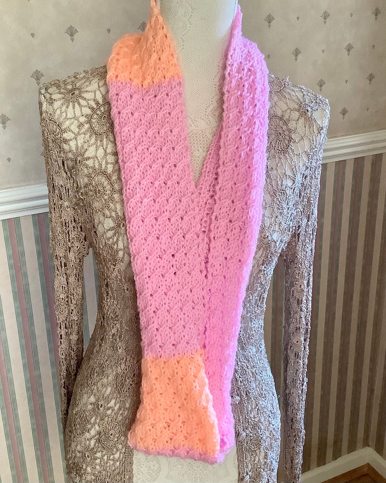 May include: A pink and peach knitted scarf with a lacy pattern. The scarf is displayed on a mannequin.