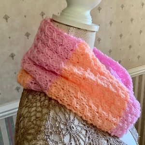 May include: A pink and peach knitted infinity scarf with a textured pattern. The scarf is displayed on a mannequin.