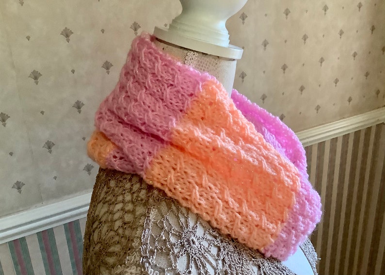 May include: A pink and peach knitted infinity scarf with a textured pattern. The scarf is displayed on a mannequin.