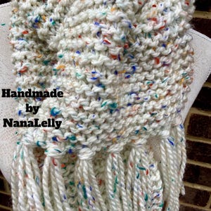 May include: A chunky knit scarf made of off-white yarn with multicolored flecks of blue, orange, and green. The scarf features a thick garter stitch texture and long, twisted fringe at the bottom. The text "Handmade by NanaLelly" is visible.