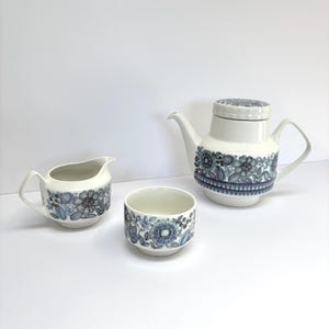 May include: A white ceramic tea set consisting of a teapot with a lid, a creamer, and a bowl. Each piece features a detailed blue and grey floral pattern with intricate line work, a curved handle on the teapot and creamer, and a smooth finish.