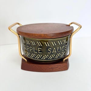 May include: A round olive green ceramic bowl with the text APPLE SAUCE embossed on the side. The bowl features a dark brown wooden lid and a matching wooden base, held together by a gold-toned metal frame with two side handles for carrying.