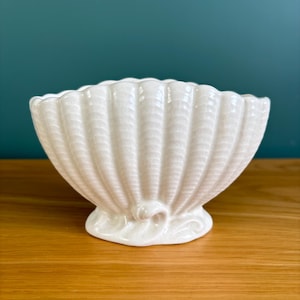 May include: A cream-coloured ceramic bowl shaped like a seashell with a scalloped rim and a ribbed exterior texture. The bowl features a glossy finish and rests on a small, decorative pedestal base with subtle swirling details at the bottom.