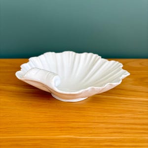 May include: A white ceramic dish shaped like a scallop shell with a smooth, glossy finish and a curved handle on one side. The bowl features deep, radiating ridges that extend from the centre to the scalloped edges, creating a textured surface.