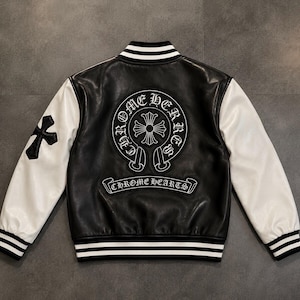 May include: A black and white varsity jacket featuring white sleeves and a black body. The back displays a large circular patch with a cross and the text CHROME HEARTS, with a banner below also reading CHROME HEARTS. A black cross is on the sleeve.