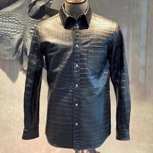 May include: A black button-down shirt made of textured leather, possibly alligator or crocodile. The shirt has a classic collar, a single chest pocket, and long sleeves. The shirt is displayed on a mannequin.