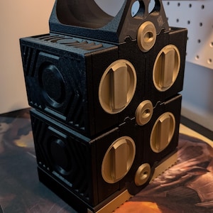 May include: A black and gold toolbox with a handle. The toolbox is made of two stacked rectangular sections with gold-colored accents and circular details. The handle is black and has a textured grip.