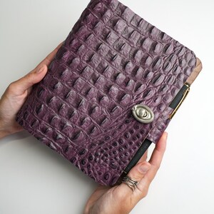 May include: A rectangular journal with a textured, deep purple cover featuring a raised, embossed crocodile skin pattern. The journal is secured with a silver-toned, oval-shaped metal twist lock closure on the front cover.