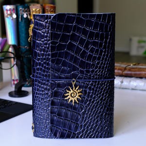 May include: A dark blue journal with a glossy crocodile skin texture. The cover is secured by a thin elastic band featuring a gold-colored sun charm with a spiral center. The journal has a vertical flap closure on the front cover.