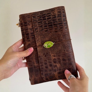 Puede incluir: Diario de cuero marrón con textura de piel de cocodrilo. El diario se asegura con una banda elástica marrón y una piedra verde ovalada decorativa. Un pequeño dije dorado cuelga de la parte superior del diario.
