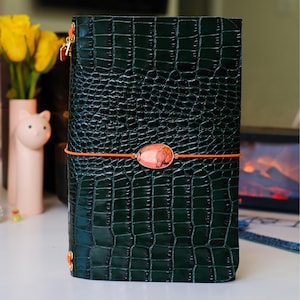 Puede incluir: Una cubierta de diario de color verde oscuro con un patrón texturizado de piel de cocodrilo. La cubierta está asegurada por una banda elástica naranja brillante con un dije central de piedra ovalada con patrones jaspeados rojos y naranjas.