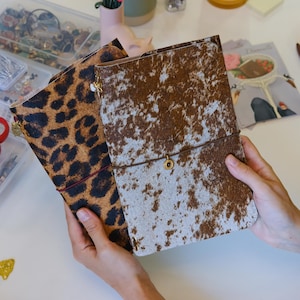 May include: Two rectangular notebooks with textured covers. One notebook features a brown and black leopard print pattern, while the other displays a brown and white cowhide pattern. Both notebooks are secured with a thin elastic band and a small charm.